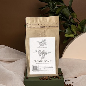 May include: A brown paper bag with a zipper closure. The bag is labeled "Southern Komforts" and features a drawing of a coffee plant. The bag contains whole bean coffee labeled "Blonde Roast" with notes of crisp buttery caramel. The bag is made in Texas and weighs 18 ounces.