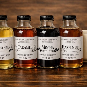 Peut inclure: Quatre bouteilles en verre de sirops de café aromatisés, dont Vanille, Caramel, Moka et Noisette, avec des bouchons noirs et des étiquettes blanches. Une tasse beige est à droite. Les bouteilles font 473 ml chacune.