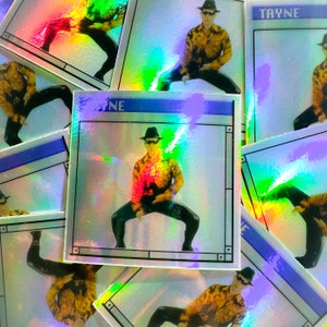 May include: A stack of iridescent stickers featuring a person in a black and gold outfit, wearing a hat and striking a pose. The stickers have a blue and white border with the word "TAYNE" printed on one of them.