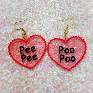 Könnte beinhalten: Ein Paar rosa herzförmige Ohrringe mit goldenen Haken. Der linke Ohrring sagt "Pee Pee" und der rechte Ohrring sagt "Poo Poo".