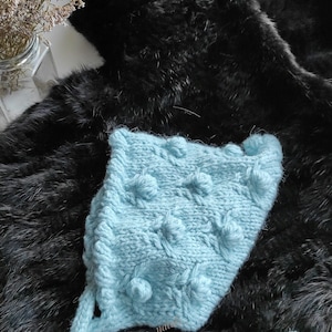 May include: A light blue knitted baby bonnet with a textured pattern and a long tie. The bonnet has a soft, fluffy texture and is made with a delicate stitch.