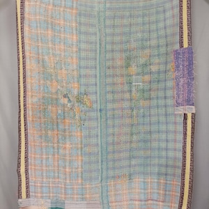 May include: A rectangular textile quilt featuring a patchwork of light blue, peach, and white plaid patterns. The fabric is stitched with visible running thread lines throughout, finished with a dark purple and yellow border along the edges.