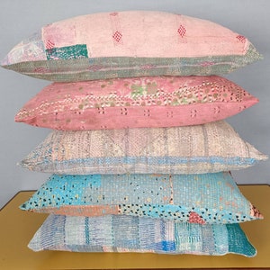 Puede incluir: Una pila de cinco cojines oblongos en varios colores y patrones. Los cojines son principalmente rosas, azules y grises, con diseños de patchwork y costuras visibles. Los cojines están apilados uno encima del otro.