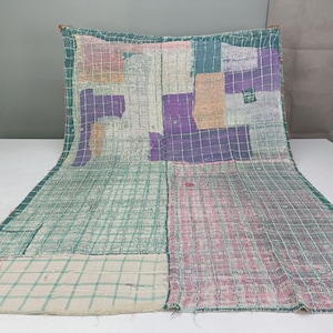 May include: A rectangular textile featuring a patchwork design with a grid pattern overlay. The fabric displays various color blocks in shades of green, purple, pink, and beige, with visible stitching throughout the surface of the material.