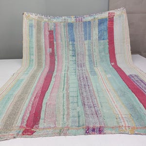 May include: A rectangular textile featuring a vertical striped pattern in shades of pink, light blue, teal, and beige. The fabric displays visible stitching throughout, creating a textured appearance with frayed edges along the top and bottom borders.