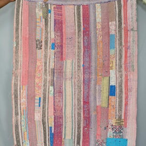 May include: A rectangular quilt constructed from vertical strips of various fabrics in shades of pink, blue, grey, and white. The textile features visible running stitch patterns throughout, with frayed edges and a patchwork design of floral and geometric prints.