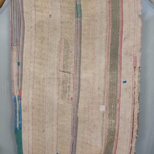 Puede incluir: Un textil rectangular que presenta un mosaico de paneles de tela en tonos blanco roto, beige y crema. La superficie está cubierta de puntadas verticales, con acentos dispersos de rayas y pequeños parches en azul, verde, rosa y gris.