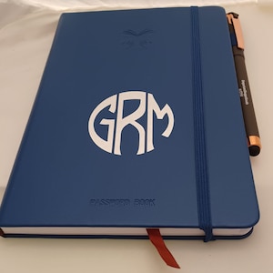 May include: A navy blue Password Book with a white monogram "GRM" and a matching elastic band closure. A rose gold and black pen rests on the side. A red ribbon bookmark is visible.