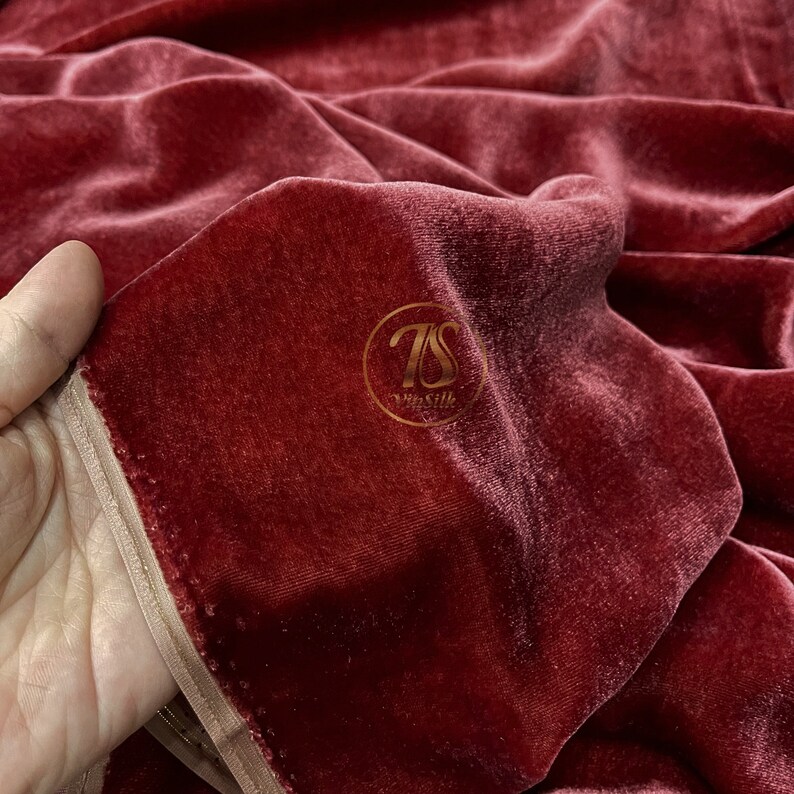 May include: Close-up of a deep red velvet fabric with a subtle sheen. The fabric is folded and draped, showcasing its soft texture. A small gold label with the text "7S The Silk" is visible in the center of the image.