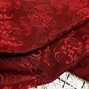 May include: Red fabric with a repeating pattern of stylized Chinese characters and floral designs. The fabric has a raised, textured surface.