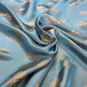 Op de afbeelding: Close-up van een turquoise stof met een bloemenpatroon. De stof heeft een zijdeachtige glans en is gedrapeerd in zachte plooien. Het bloemmotief is in een contrasterende goudkleur, wat een luxueuze uitstraling creëert.