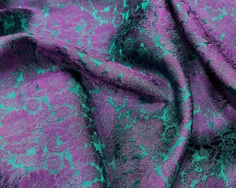 Pode incluir: Tecido roxo com um padrão floral turquesa. O tecido é texturizado e tem um brilho sutil.