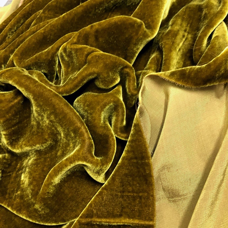 May include: Close-up of a rich, olive green velvet fabric with a smooth, silky texture. The fabric is draped and folded, showcasing its luxurious drape and soft sheen.