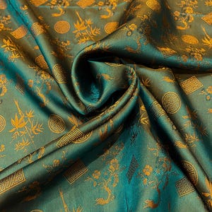 Könnte beinhalten: Smaragdgrüner Stoff mit goldenem Blumen- und geometrischem Muster. Der Stoff ist drapiert und zeigt den Glanz und die Textur des Materials. Das Design umfasst stilisierte Bäume, Blumen sowie kreisförmige und rechteckige Motive.