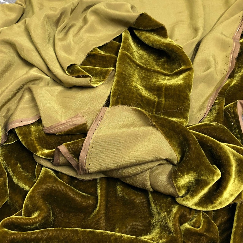 May include: A close-up of a pile of olive green velvet fabric. The fabric has a soft, luxurious feel and a subtle sheen.