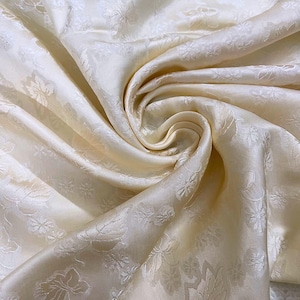 Pode incluir: Tecido de cor creme com um padrão floral em relevo sutil. O tecido é macio e luxuoso, com um leve brilho.