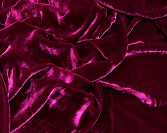 Könnte beinhalten: Nahaufnahme eines tief magentafarbenen Samtstoffes mit weicher, luxuriöser Textur. Der Stoff ist drapiert und gefaltet und erzeugt einen dramatischen und eleganten Look.