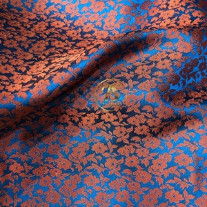 May include: A close-up of a blue fabric with a repeating pattern of small orange flowers. The fabric has a subtle sheen and a textured surface.