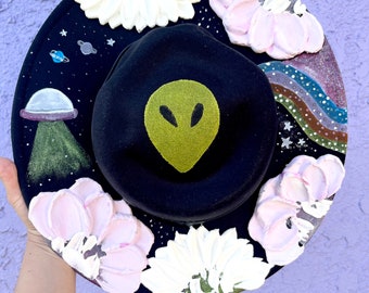 May include: A black hat with a green alien face painted on the front. The hat is decorated with white and pink flowers and a galaxy scene with stars and a rainbow.