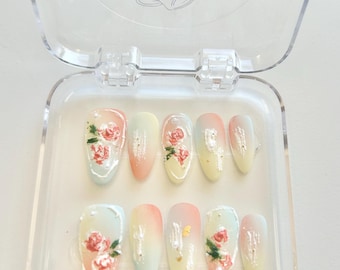 Può includere: Un set di dieci unghie finte ovali in una custodia di plastica trasparente. Ogni unghia presenta un design sfumato in colori pastello, adornato con rose rosse dipinte a mano e dettagli in foglia d'oro. La custodia ha il testo "BY DIDY".