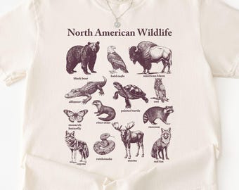 May include: Cream-colored t-shirt featuring a graphic of North American wildlife. The design includes illustrations of a black bear, bald eagle, bison, alligator, turtle, and other animals. The words "North American Wildlife" are printed above the illustrations.