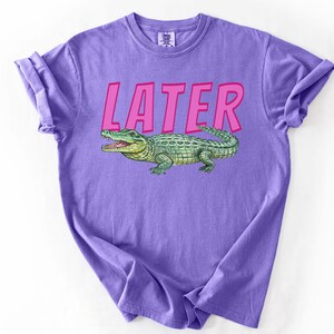 May include: A lilac t-shirt featuring the word "LATER" in pink lettering above a green and yellow alligator illustration. The t-shirt is folded with rolled-up sleeves.