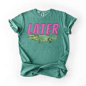 May include: Teal t-shirt featuring the word "LATER" in pink block letters above a green alligator illustration. The alligator has an open mouth. The shirt is folded.