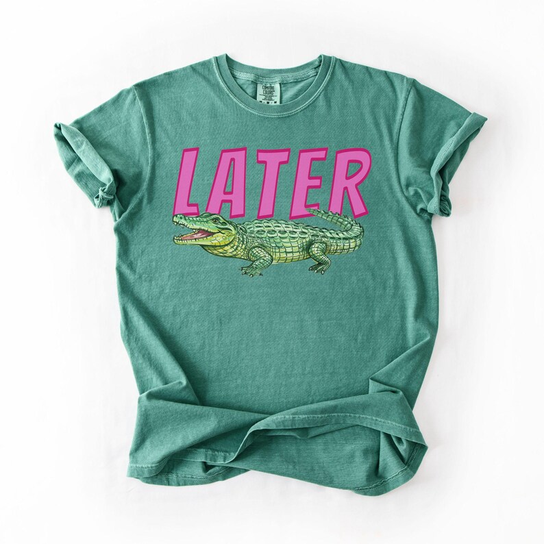 May include: Teal t-shirt featuring the word "LATER" in pink block letters above a green alligator illustration. The alligator has an open mouth. The shirt is folded.