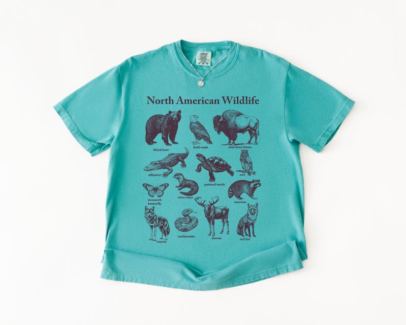 Pu&ograve; includere: Una maglietta turchese con una grafica della fauna selvatica nordamericana. Il design include illustrazioni di orso nero, aquila calva, bisonte, alligatore, tartaruga, gufo, farfalla, lontra di fiume, procione, lupo, serpente, alce e volpe rossa.