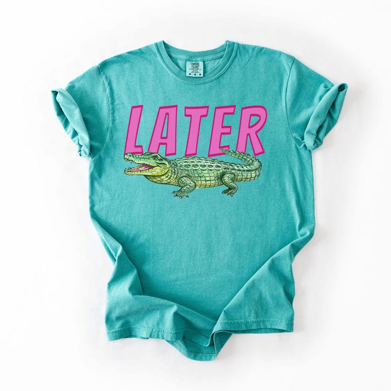May include: Teal t-shirt featuring the word "LATER" in pink block letters above a detailed illustration of a green alligator. The alligator has yellow accents and is facing left with its mouth open.