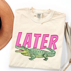 May include: Cream-coloured t-shirt featuring the word "LATER" in bright pink letters and a green alligator graphic. The alligator has open jaws and a textured body. A gold necklace and a brown hat are visible.