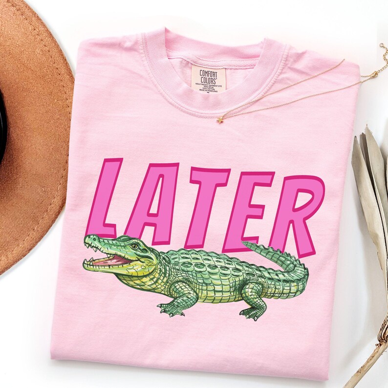 May include: A pale pink t-shirt featuring the word "LATER" in bold pink letters and a green alligator illustration. The alligator has yellow highlights and an open mouth. The shirt is folded and has a Comfort Colors label.