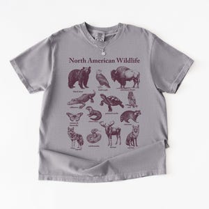 Pu&ograve; includere: Una maglietta grigia con una grafica bordeaux raffigurante la fauna selvatica nordamericana. Il design include illustrazioni di un orso, un'aquila, un bisonte, un alligatore, una tartaruga e altri animali. Le parole "North American Wildlife" sono stampate sopra le illustrazioni.