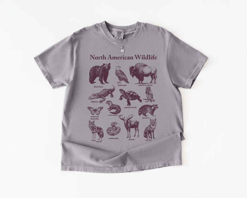 Pu&ograve; includere: Una maglietta grigia con una grafica bordeaux raffigurante la fauna selvatica nordamericana. Il design include illustrazioni di un orso, un'aquila, un bisonte, un alligatore, una tartaruga e altri animali. Le parole "North American Wildlife" sono stampate sopra le illustrazioni.
