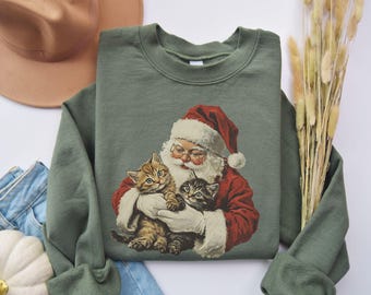 May include: Sage green sweatshirt featuring a vintage illustration of Santa Claus holding two kittens. Santa is wearing a red suit and hat with white trim. The kittens have brown and gray fur. The sweatshirt has a crew neck and long sleeves.