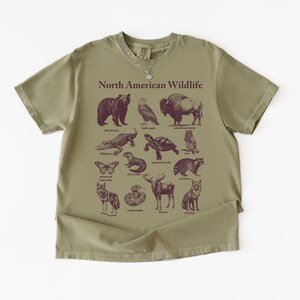 Pu&ograve; includere: T-shirt verde oliva con la scritta "North American Wildlife" e illustrazioni di vari animali. Gli animali includono un orso, un'aquila, un bisonte, un alligatore, una tartaruga, un gufo, una farfalla, una lontra, un procione, un lupo, un alce, un serpente e una volpe.