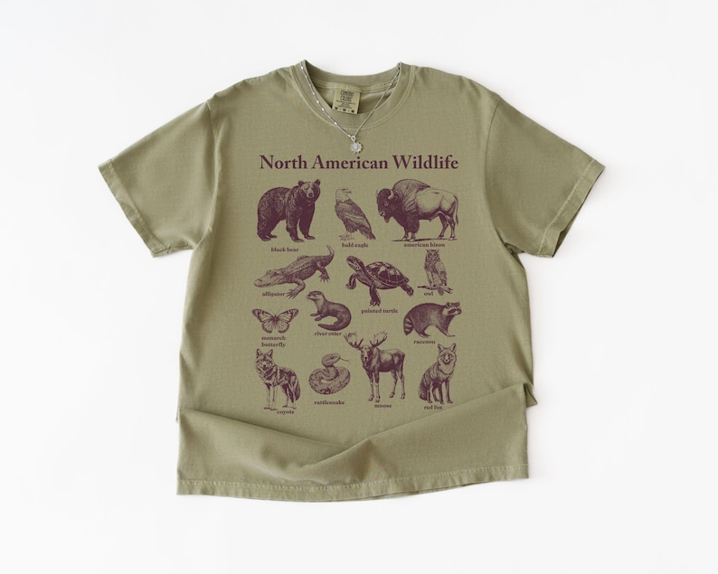 Pu&ograve; includere: T-shirt verde oliva con la scritta "North American Wildlife" e illustrazioni di vari animali. Gli animali includono un orso, un'aquila, un bisonte, un alligatore, una tartaruga, un gufo, una farfalla, una lontra, un procione, un lupo, un alce, un serpente e una volpe.