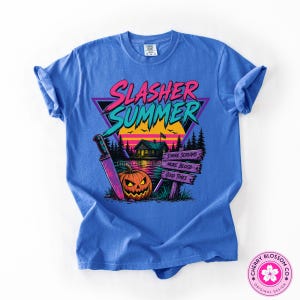 Puede incluir: Una camiseta azul con un estampado gr&aacute;fico que incluye el texto Slasher Summer. El dise&ntilde;o muestra una caba&ntilde;a, una calabaza, un cuchillo grande y un poste de madera con el texto S'more Screams, More Blood y Good Times al atardecer.