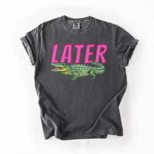 May include: Dark grey t-shirt featuring the word "LATER" in pink above a green alligator illustration. The alligator has yellow accents and is facing left. The shirt has rolled-up sleeves.