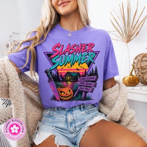 Puede incluir: Una camiseta de color morado claro con un estampado gr&aacute;fico con el texto SLASHER SUMMER en rosa y turquesa. El dise&ntilde;o incluye una caba&ntilde;a, una puesta de sol, un cuchillo grande, una calabaza y un letrero de madera con S'MORE SCREAMS, MORE BLOOD, GOOD TIMES.