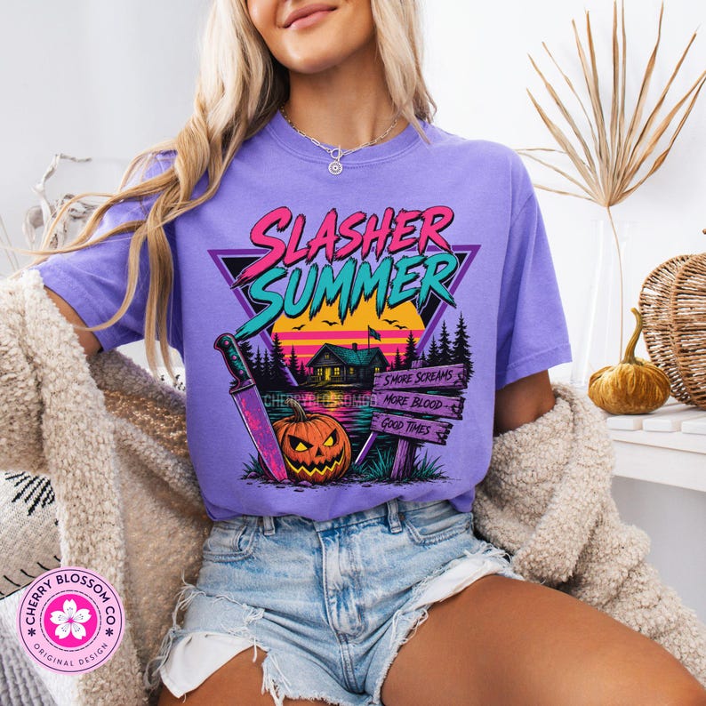 Puede incluir: Una camiseta de color morado claro con un estampado gr&aacute;fico con el texto SLASHER SUMMER en rosa y turquesa. El dise&ntilde;o incluye una caba&ntilde;a, una puesta de sol, un cuchillo grande, una calabaza y un letrero de madera con S'MORE SCREAMS, MORE BLOOD, GOOD TIMES.