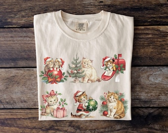 May include: Cream-colored t-shirt featuring six Christmas-themed kitten illustrations. The kittens are depicted with Santa hats, ornaments, and gifts. The shirt has a Comfort Colors tag.