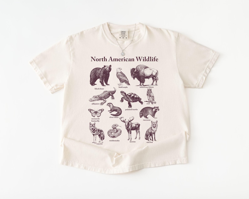 Pu&ograve; includere: T-shirt color crema con una grafica della fauna selvatica nordamericana. Il design include illustrazioni di orso nero, aquila calva, bisonte, alligatore, tartaruga e altri animali. Le parole "North American Wildlife" sono stampate sopra le illustrazioni.