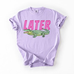 May include: A lilac t-shirt featuring the word "LATER" in pink, above a green and yellow alligator illustration. The t-shirt is made of soft material and has rolled-up sleeves.