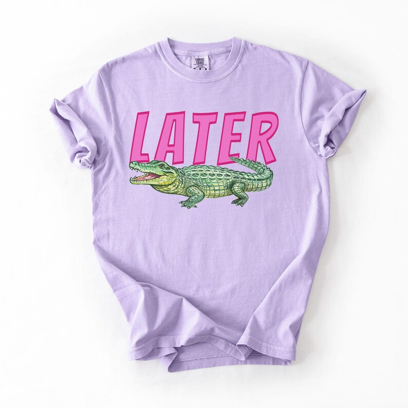 May include: A lilac t-shirt featuring the word "LATER" in pink, above a green and yellow alligator illustration. The t-shirt is made of soft material and has rolled-up sleeves.