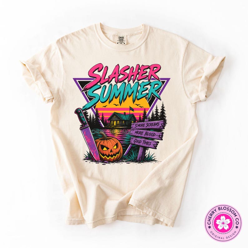 Puede incluir: Una camiseta color crema con un gr&aacute;fico que incluye el texto SLASHER SUMMER en rosa y turquesa. El dise&ntilde;o muestra un cuchillo grande, una calabaza, una caba&ntilde;a junto a un lago y un letrero de madera con el texto SMORE SCREAMS, MORE BLOOD, GOOD TIMES.