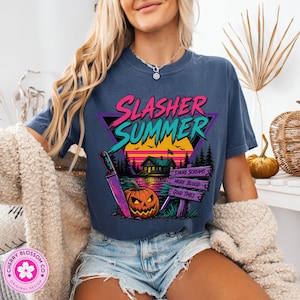 Puede incluir: Una camiseta azul oscuro con un estampado gr&aacute;fico que incluye el texto "SLASHER SUMMER" en rosa y turquesa. El dise&ntilde;o muestra una caba&ntilde;a, un atardecer, una calabaza, un cuchillo grande y un letrero con "SMORE SCREAMS", "MORE BLOOD", "GOOD TIMES".