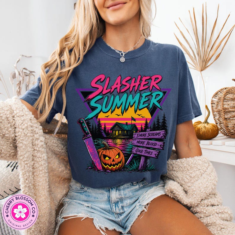 Puede incluir: Una camiseta azul oscuro con un estampado gr&aacute;fico que incluye el texto "SLASHER SUMMER" en rosa y turquesa. El dise&ntilde;o muestra una caba&ntilde;a, un atardecer, una calabaza, un cuchillo grande y un letrero con "SMORE SCREAMS", "MORE BLOOD", "GOOD TIMES".