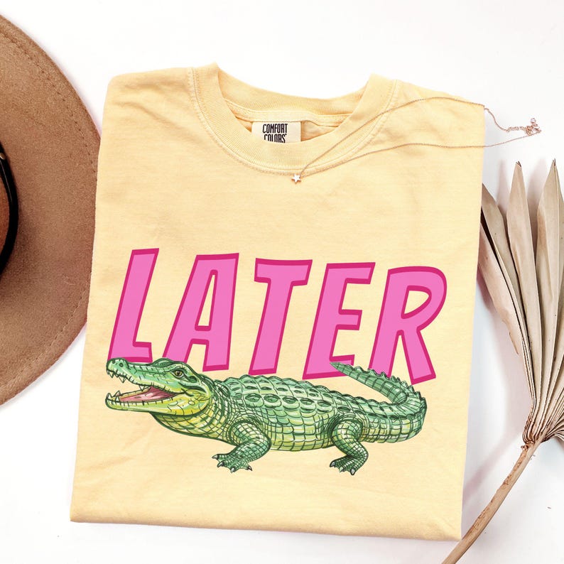 May include: A pale yellow t-shirt featuring the word "LATER" in pink, with a green alligator illustration. The shirt is from "Comfort Colors". A gold necklace with a star pendant is on the shirt.