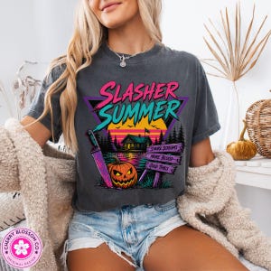 Puede incluir: Una camiseta gris carb&oacute;n de manga corta con un estampado gr&aacute;fico que incluye el texto Slasher Summer, una caba&ntilde;a, una calabaza, un cuchillo grande y un letrero con las palabras More Screams, More Blood y Good Times en colores vivos.